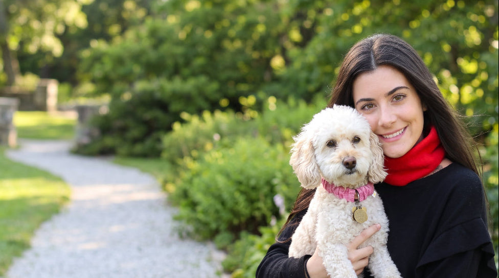 A woman with long dark hair smiling while holding a small white poodle wearing a sandy collar wrap in an outdoor garden setting with a gravel path and green foliage in the background.